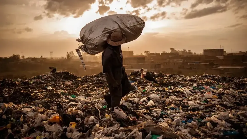 A waste picker carries a large sack across a dumpsite at sunset, with the city behind.
