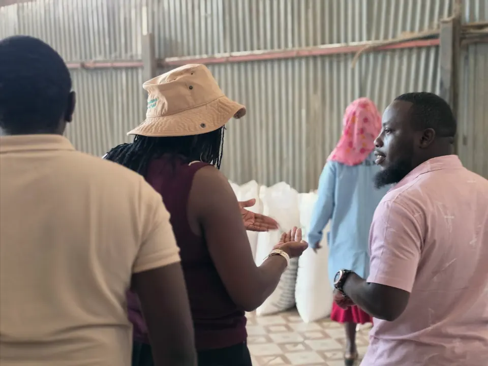 Lucy Nyoike and Zecharia Mwangi of ENKOP in discussion with partners in a storeroom stacked with white sacks.