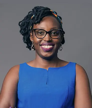 Portrait of Lucy Nyoike, smiling, in glasses and a blue sleeveless top, against a grey studio background.