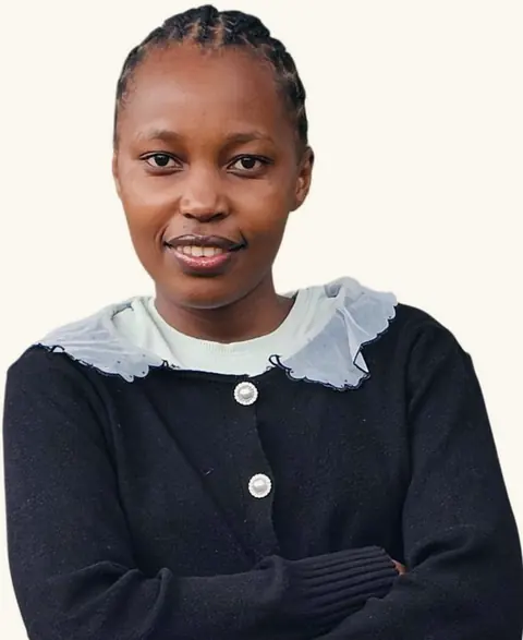 Portrait of Mercy Wanjiku, smiling with arms folded, in a black cardigan with a sheer white collar, against a white background.
