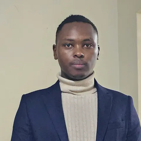Portrait of Zecharia Mwangi in a navy blazer over a cream cable-knit roll-neck, against a plain wall.